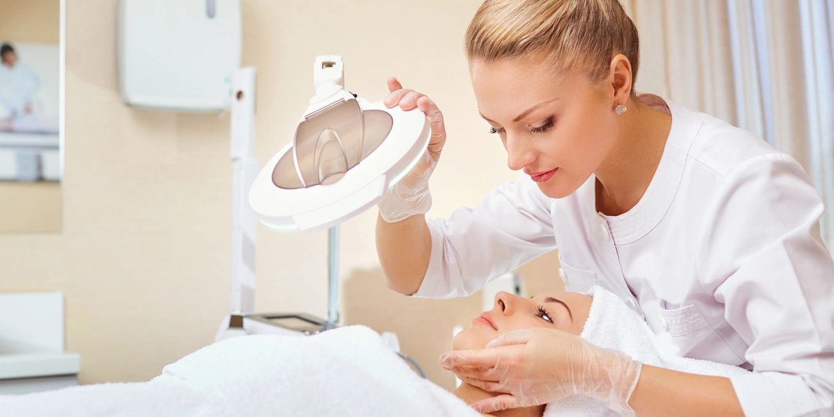 A cosmetologist examining a patient's face under a magnifying lamp.